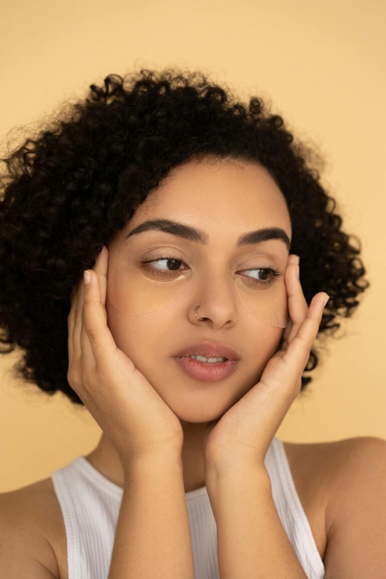 A woman in a white tank top gently caressing her face with under-eye masks.