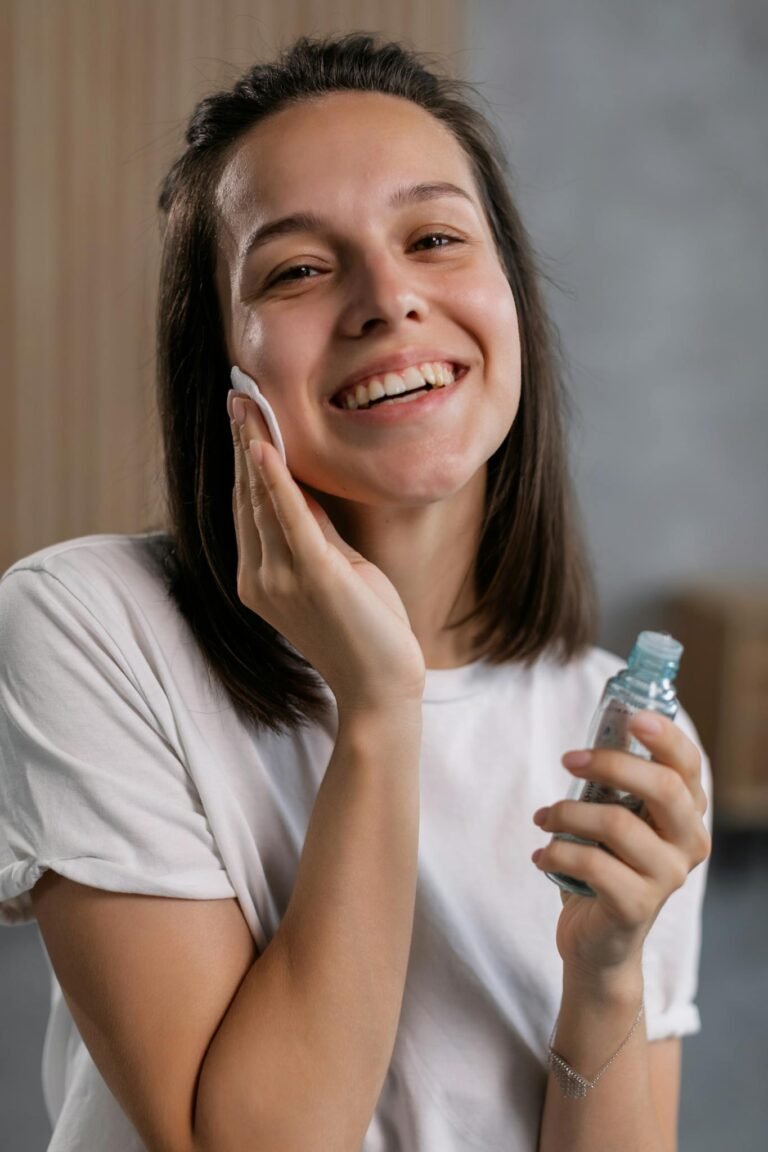 A woman smiles while applying a skincare product indoors, emphasizing beauty and facial care.