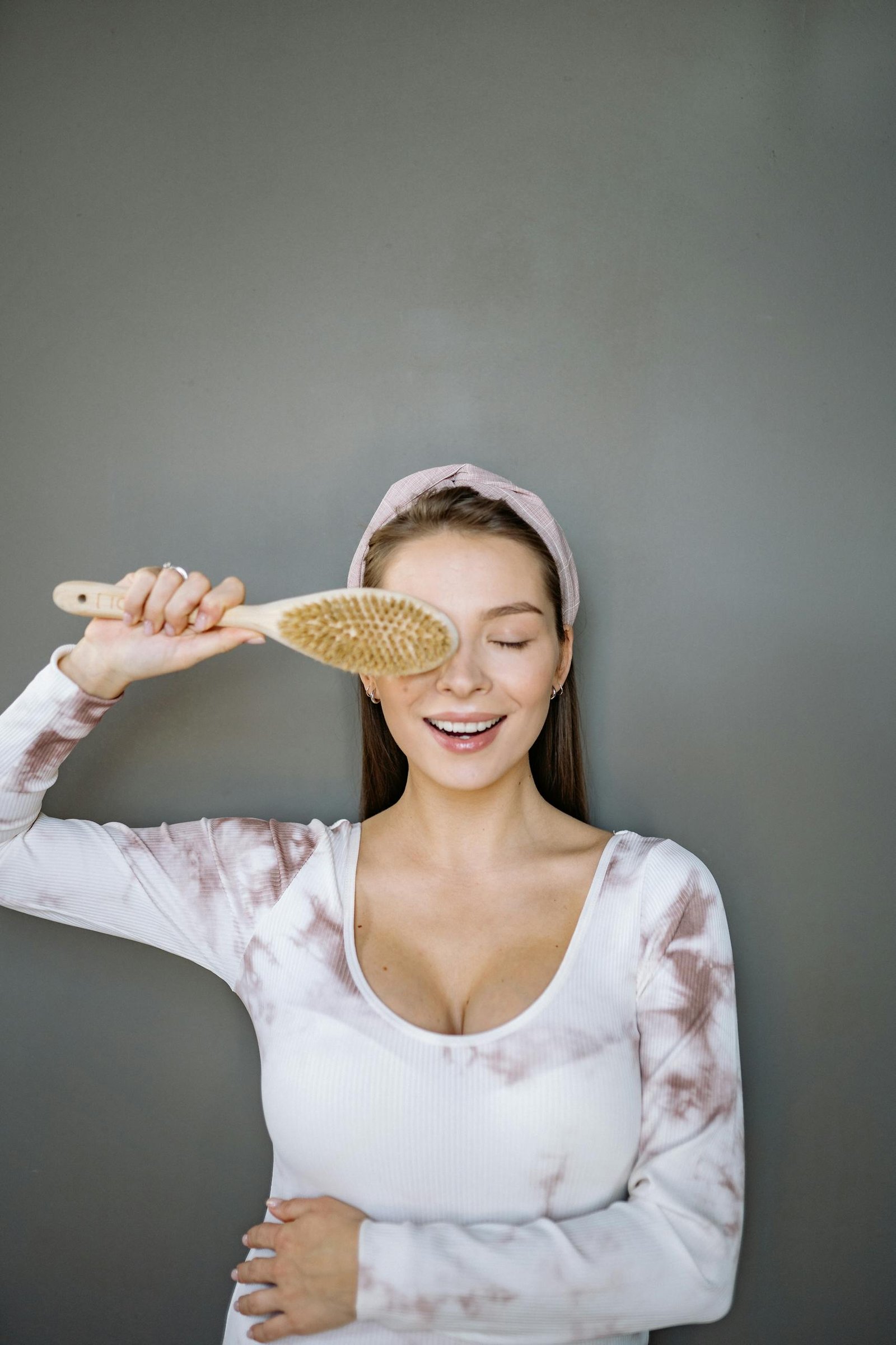 Caucasian woman smiling with eyes closed while holding a natural brush against a minimalist background.