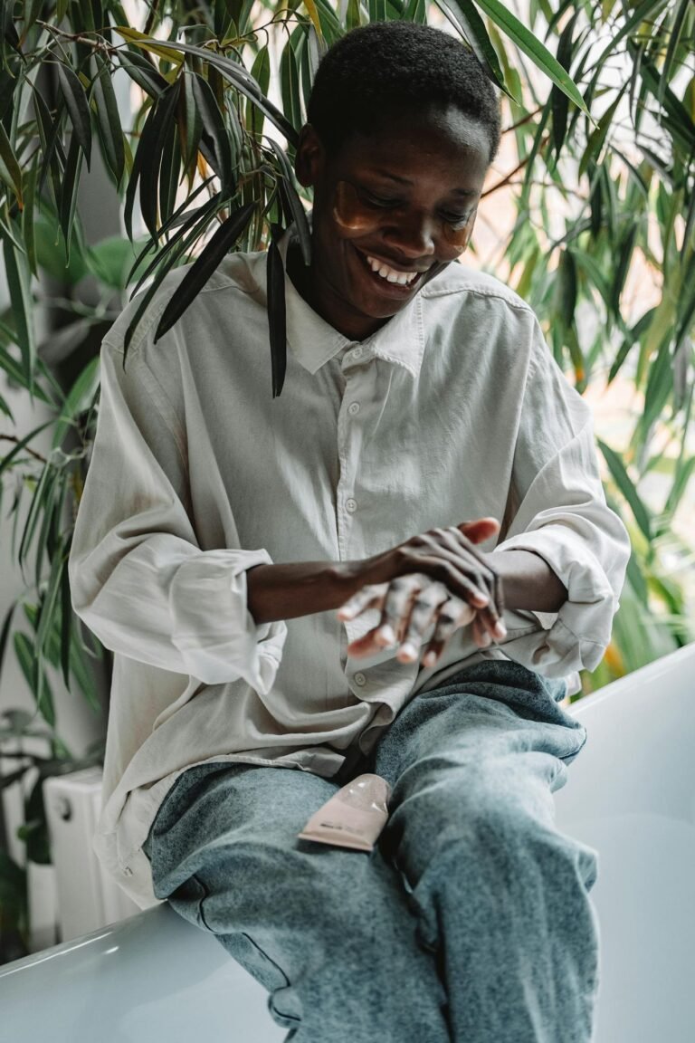 Happy woman applying lotion while sitting indoors, surrounded by plants, embracing skincare routine.